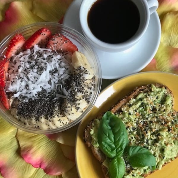 Acai bowl with strawberries, coconut, and chia seeds, avocado toast, and coffee on a yellow surface.