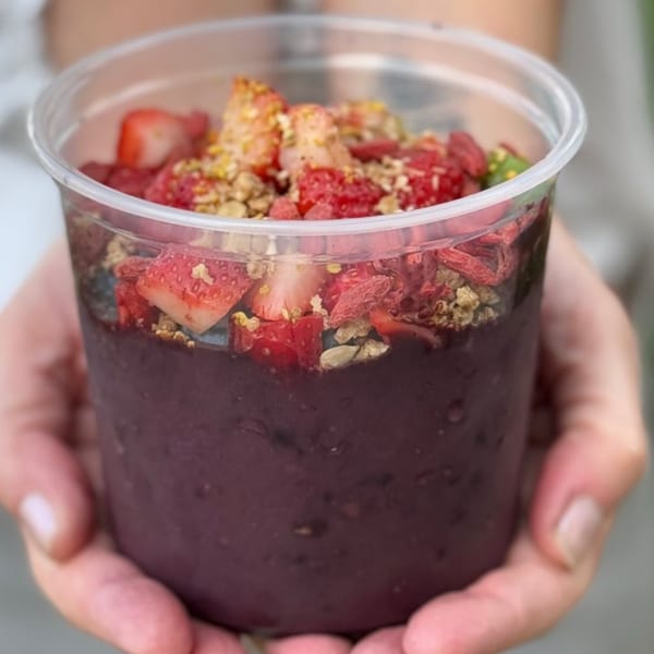 Hands holding a clear cup filled with a purple acai bowl topped with strawberries, goji berries, and granola.