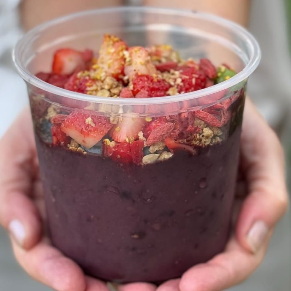 Hands holding a clear cup filled with a purple acai bowl topped with strawberries, goji berries, and granola.