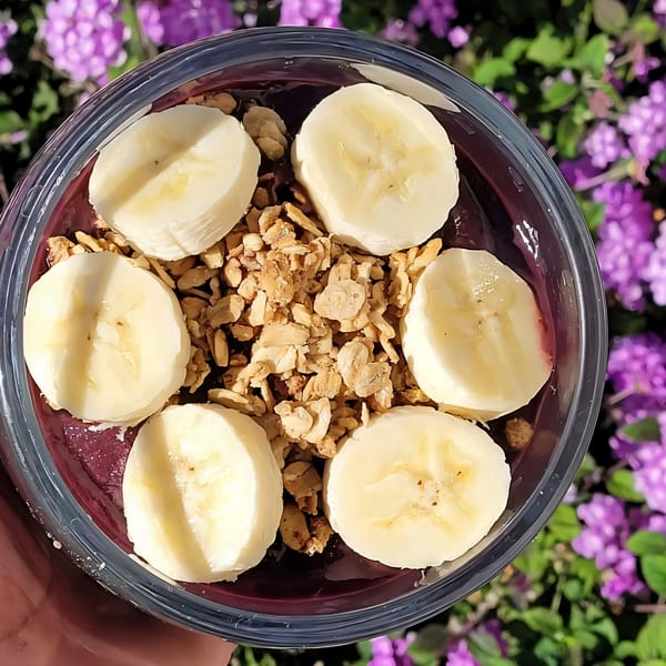 Overhead view of an acai bowl with banana slices and granola, against a purple floral background.