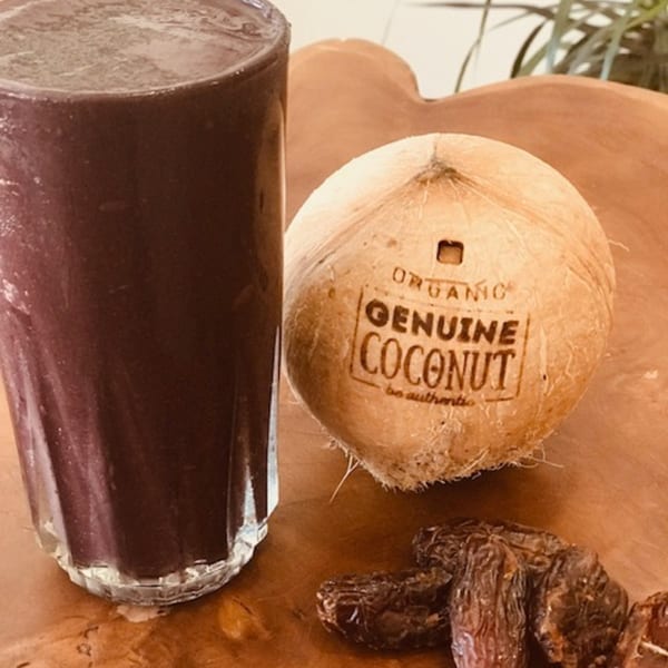 A dark purple acai smoothie, a whole coconut, and dried dates on a wooden surface.
