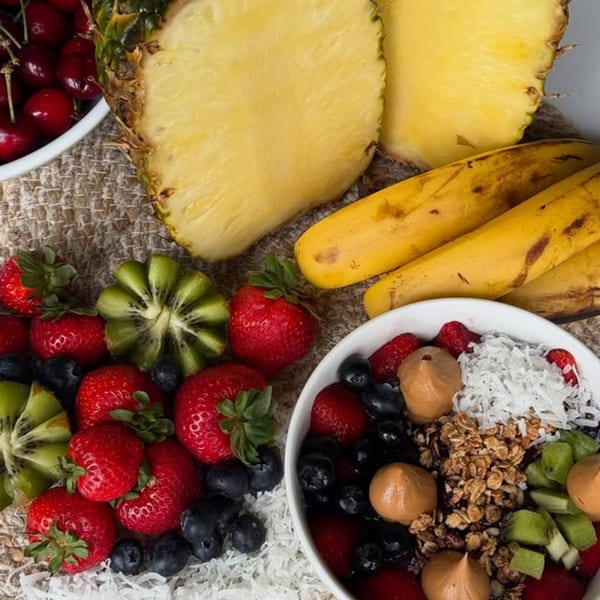 An acai bowl with granola, coconut, peanut butter, and fresh fruit, surrounded by whole fruits.