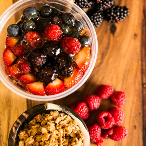 A plastic cup of acai bowl with mixed berries, granola, and fresh raspberries on a wooden table.