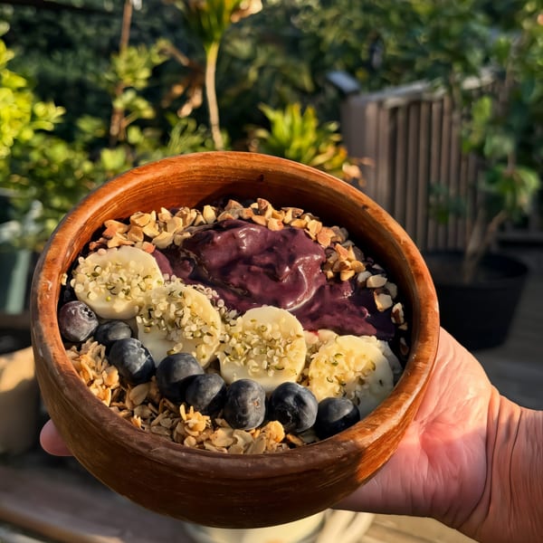 A hand holds a wooden bowl filled with an acai bowl topped with bananas, blueberries, granola, and nuts.